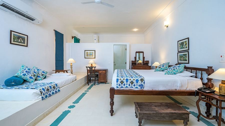 Room with two beds, patterned pillows and runners, furniture including desk, tables, and a trunk, artwork, wall-mounted air conditioner, and patterned floor tiles - Tijara Fort-Palace - 19th Century, Alwar.