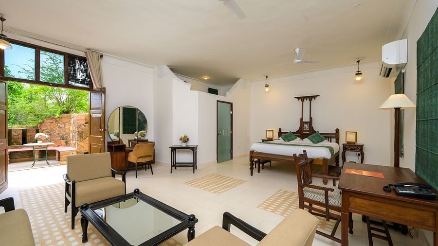 A spacious room featuring a wooden four-poster bed, a seating area with beige chairs, a dark wood desk, and an open doorway leading to an outdoor space, offering a blend of indoor and outdoor living - Tijara Fort-Palace - 19th Century, Alwar.