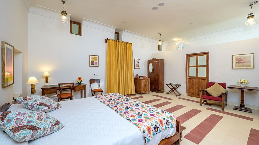 Room with bed, patterned pillows and runner, furniture including desk, chair, wardrobe, and tables, artwork, decorative ceiling lights, and patterned floor tiles - Tijara Fort-Palace - 19th Century, Alwar.