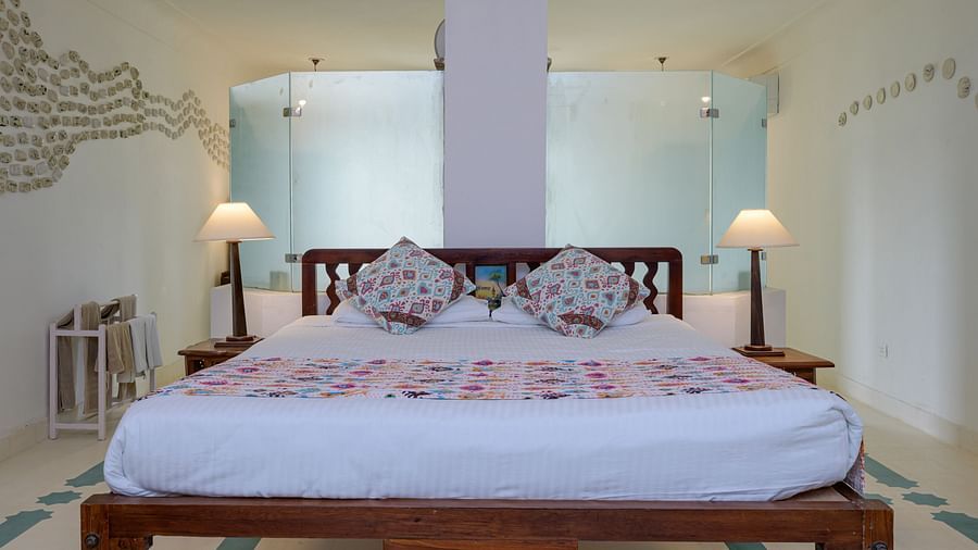 Bedroom with a wooden bed frame, white linens, patterned pillows and bedspread, bedside tables with lamps, and a frosted glass partition at Tijara Fort-Palace - 19th Century, Alwar.