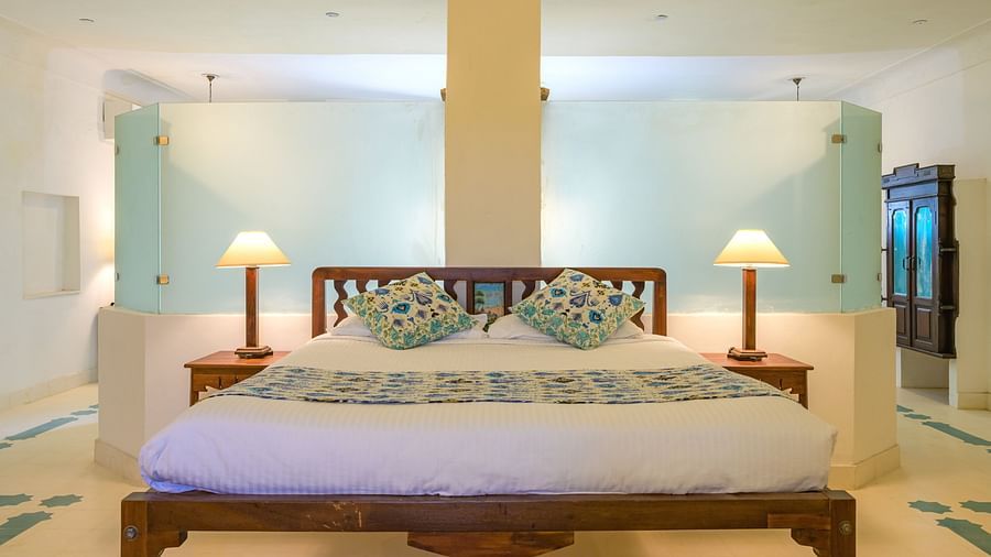 A bedroom with a wooden bed frame, white linens, patterned pillows and bedspread, bedside tables with lamps, and frosted glass partitions on either side at Tijara Fort-Palace - 19th Century, Alwar.