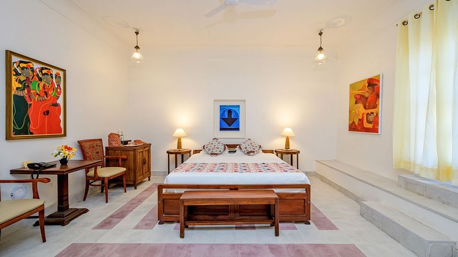 Room with bed, patterned pillows and runner, furniture including desk and folding table, artwork, decorative ceiling lights, patterned floor tiles, and a window with stained glass - Tijara Fort-Palace - 19th Century, Alwar.