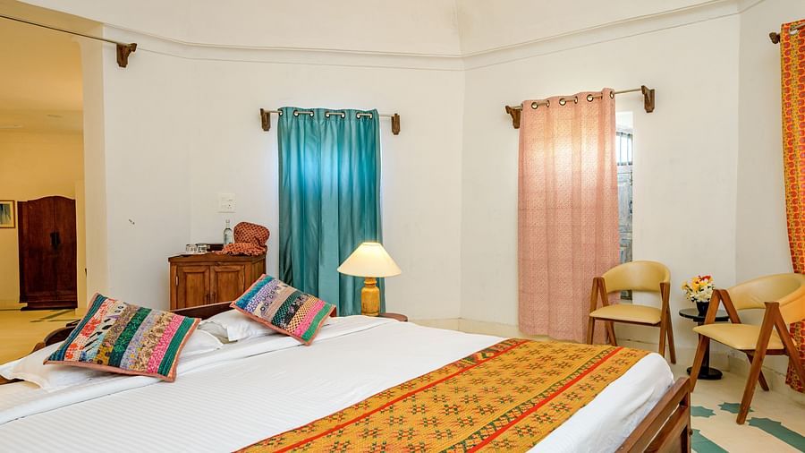 Room with bed, patterned pillows, curtains, windows, chairs, and a lamp - Tijara Fort-Palace - 19th Century, Alwar.