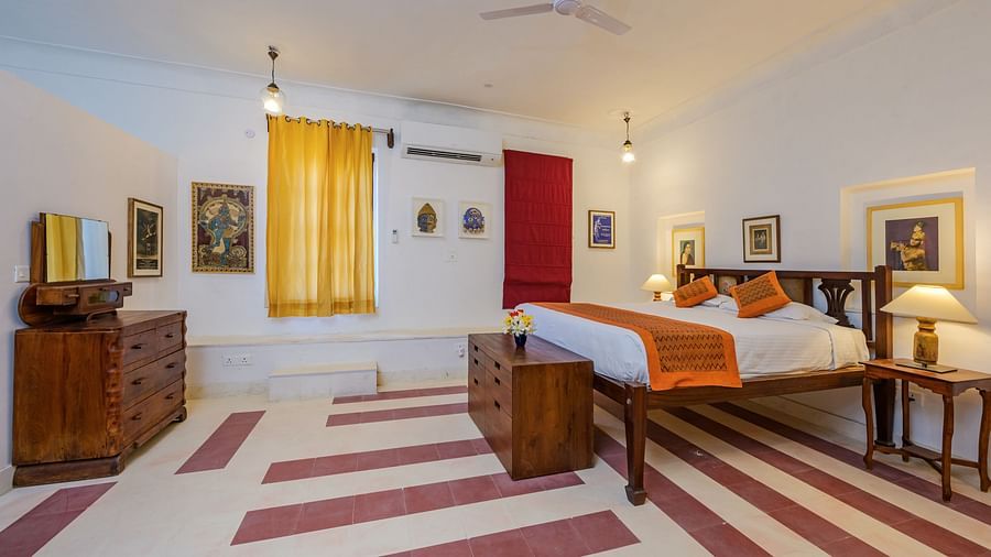 Room with bed, patterned pillows and runner, furniture including dresser, tables, and trunk, artwork, yellow and red curtains, decorative ceiling lights, and patterned floor tiles - Tijara Fort-Palace - 19th Century, Alwar.