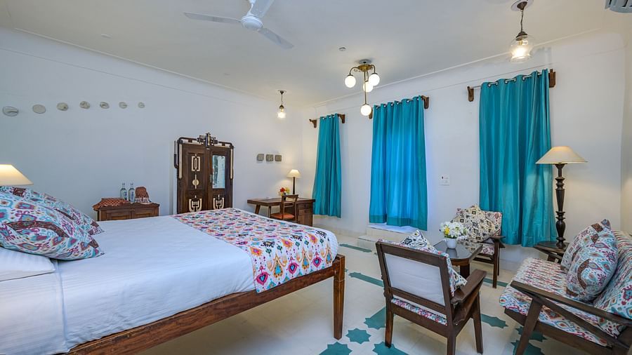 Bedroom with a bed, patterned bedspread and pillows, wooden furniture including a wardrobe and desk, two chairs with a small table, and windows with curtains at Tijara Fort-Palace - 19th Century, Alwar.