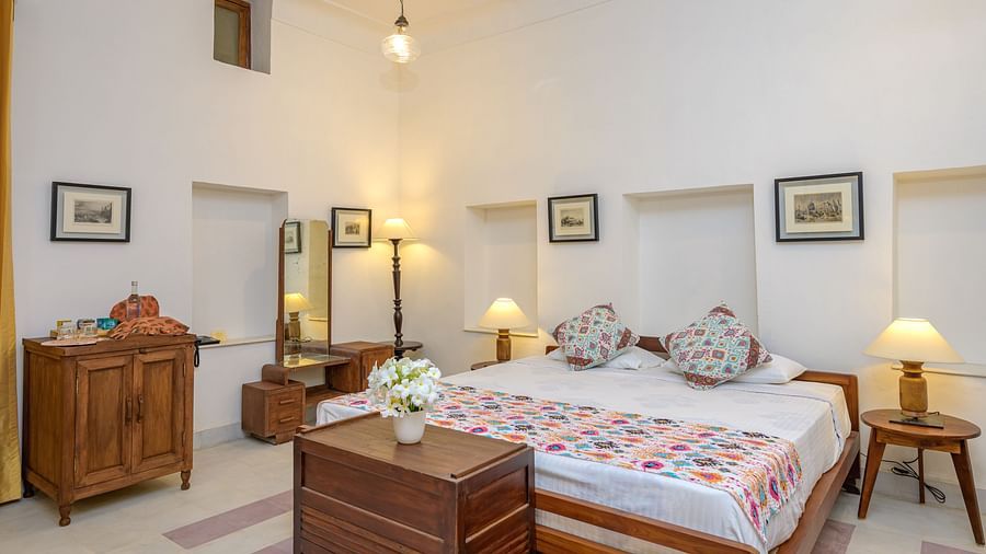 A bedroom with a bed featuring patterned bedding, wooden furniture including a dresser, a dressing table with a mirror, and bedside tables with lamps at Tijara Fort-Palace - 19th Century, Alwar.