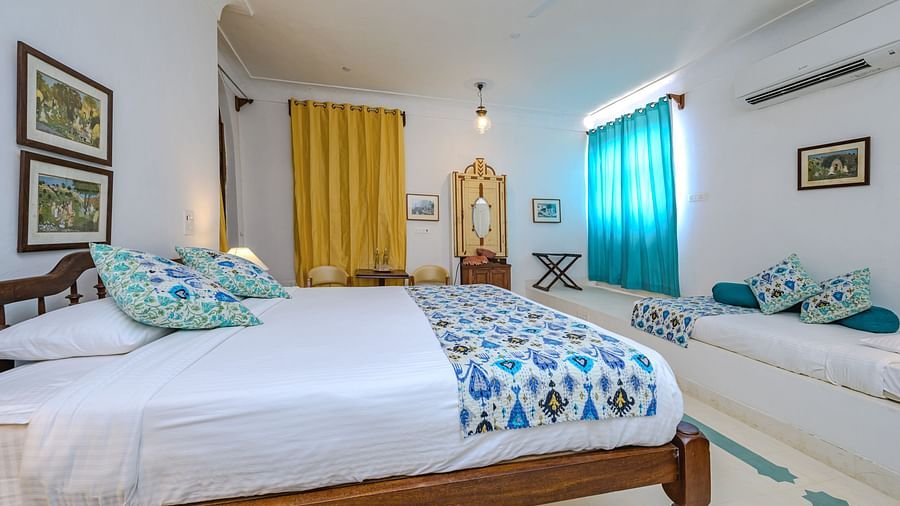 Room with bed, patterned pillows and runner, small daybed with patterned pillows, artwork, yellow and blue curtains, decorative mirror, wall-mounted air conditioner, and patterned floor tiles - Tijara Fort-Palace - 19th Century, Alwar.
