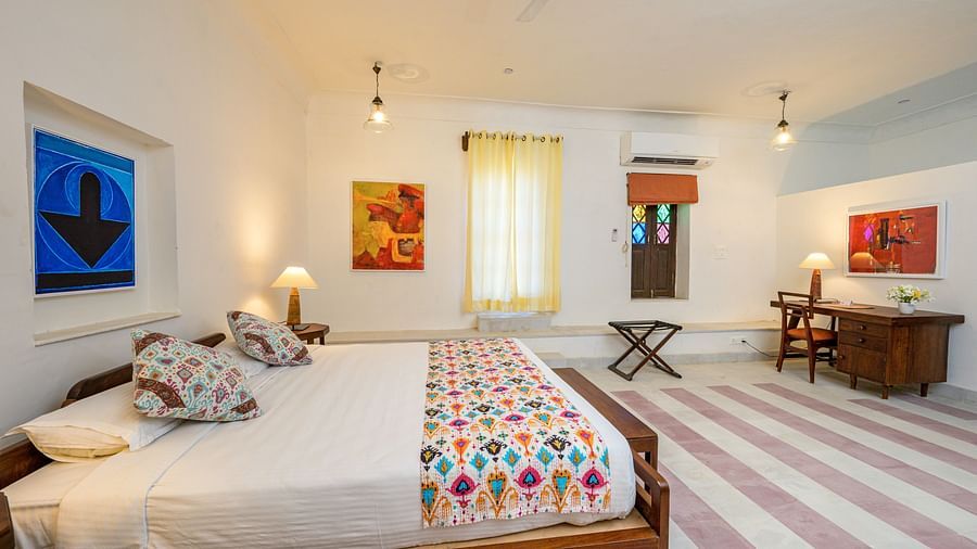 Room with bed, patterned pillows and runner, furniture including desk and folding table, artwork, decorative ceiling lights, patterned floor tiles, and a window with stained glass - Tijara Fort-Palace - 19th Century, Alwar.