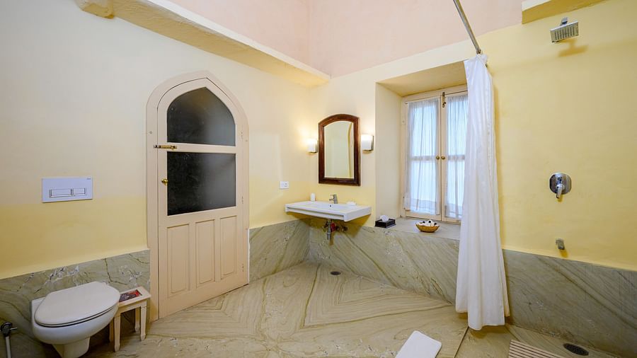 A bathroom with a sink, mirror, toilet, door, and shower curtain in Tijara Fort-Palace - 19th Century, Alwar.