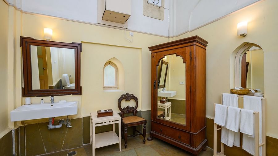 A bathroom with a sink, mirror, wardrobe, chair, and towel rack with towels in Tijara Fort-Palace - 19th Century, Alwar.