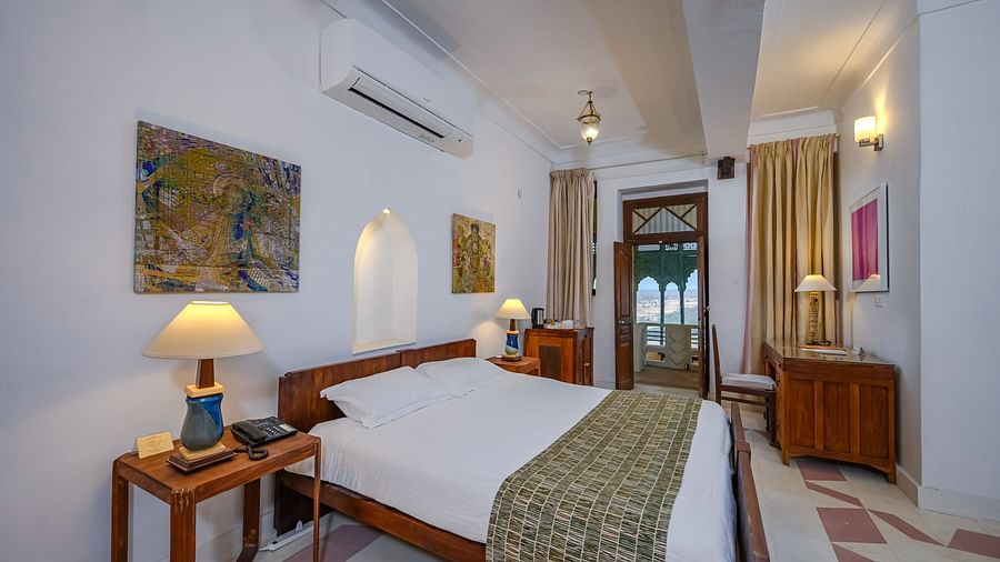 A bed with white linens and a patterned throw is in a bedroom at Tijara Fort-Palace - 19th Century, Alwar, which also features bedside tables with lamps, artwork on the walls, and a glimpse into an adjacent area.