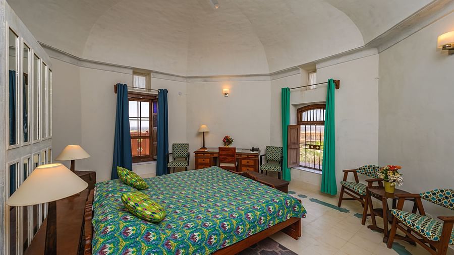 Room with bed, patterned bedding and pillows, furniture including desk, chairs, and tables, windows with curtains, decorative wall lights, and unique floor pattern - Tijara Fort-Palace - 19th Century, Alwar.