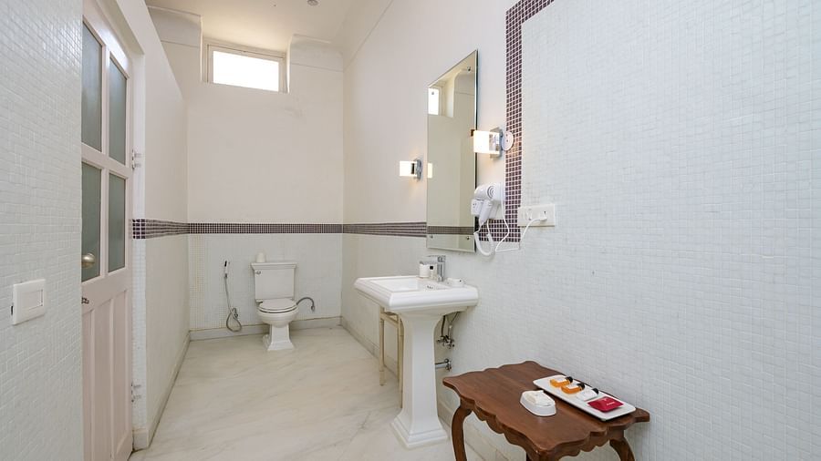 A bathroom with a toilet, a pedestal sink with a mirror above it, and a small table with toiletries at Tijara Fort-Palace - 19th Century, Alwar.
