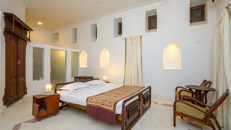 Room with bed, wooden frame, patterned runner, furniture including a wardrobe and chairs, curtains, and arched alcoves in the wall - Tijara Fort-Palace - 19th Century, Alwar.