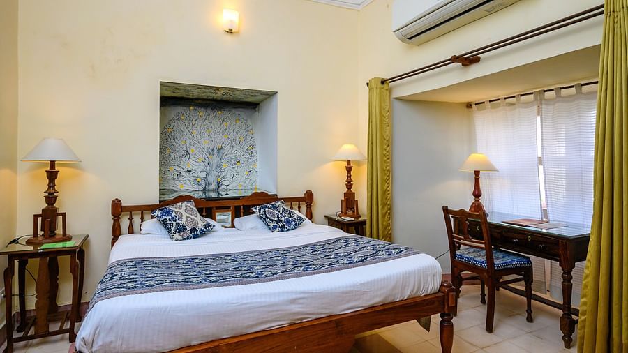 Room with bed, patterned runner and pillows, furniture including tables and desk, lamps, curtains, wall-mounted air conditioner, and window with patterned drapes - Tijara Fort-Palace - 19th Century, Alwar.