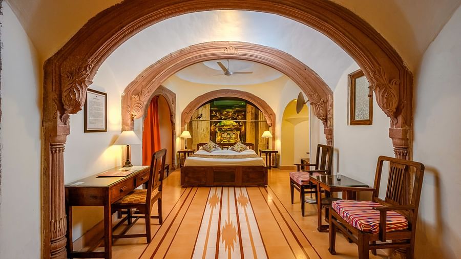 A hotel hallway with arched openings leading to a bedroom with a bed and other furnishings, as well as a desk and chairs along the hallway at Tijara Fort-Palace - 19th Century, Alwar.