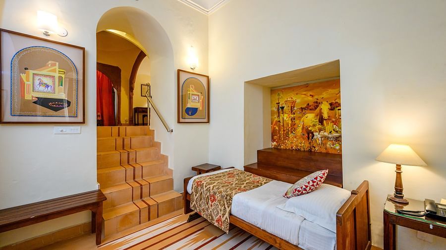 A hotel room with a single bed featuring patterned bedding, a bedside table with a lamp, a raised platform with artwork, stairs leading to another level, and framed pictures on the walls at Tijara Fort-Palace - 19th Century, Alwar.