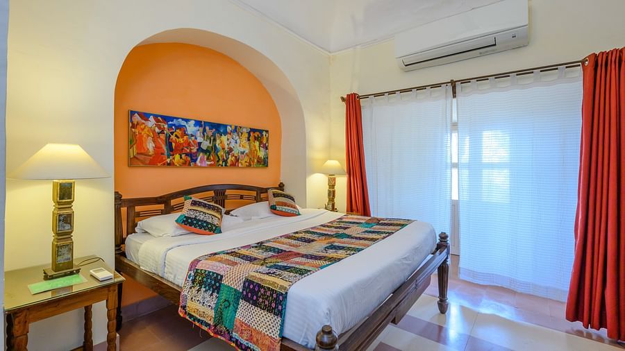 Bedroom with a bed featuring patterned bedding, bedside tables with lamps, an alcove with artwork, and curtains at Tijara Fort-Palace, Alwar.