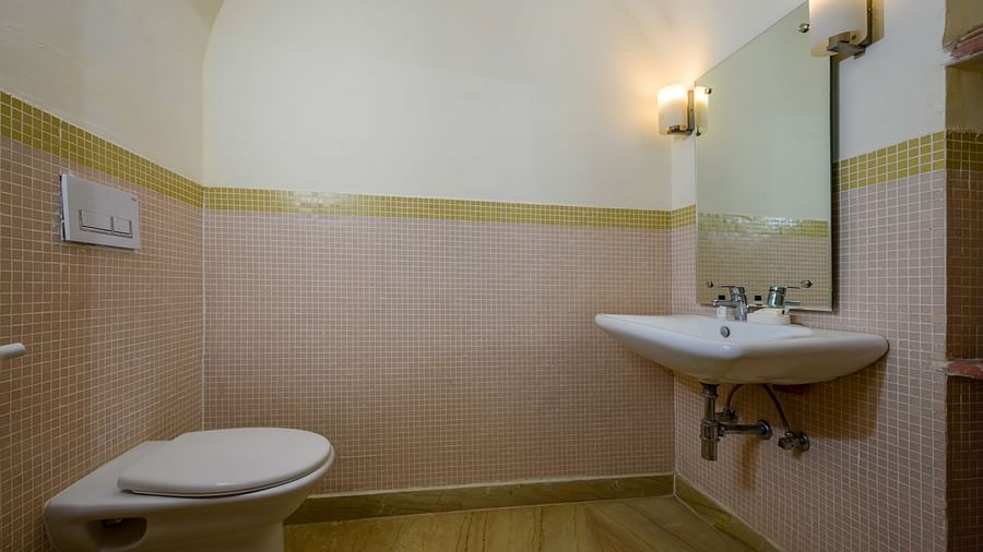 Bathroom with a toilet, a sink with a mirror and lights, and tiled walls at Tijara Fort-Palace, Alwar.