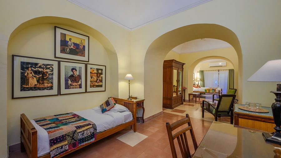 Room with single bed, patterned runner and pillow, furniture including desk, chair, tables, and wardrobe, framed artwork, arched doorways, and patterned floor tiles - Tijara Fort-Palace - 19th Century, Alwar.