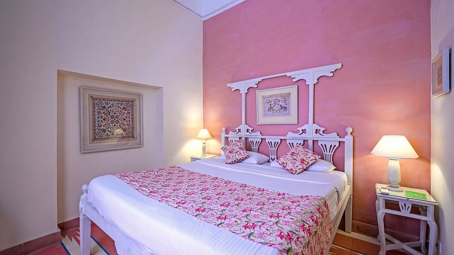 A bedroom with a bed featuring floral bedding, a decorative headboard, a bedside table with a lamp, and framed artwork on the walls at Tijara Fort-Palace - 19th Century, Alwar.