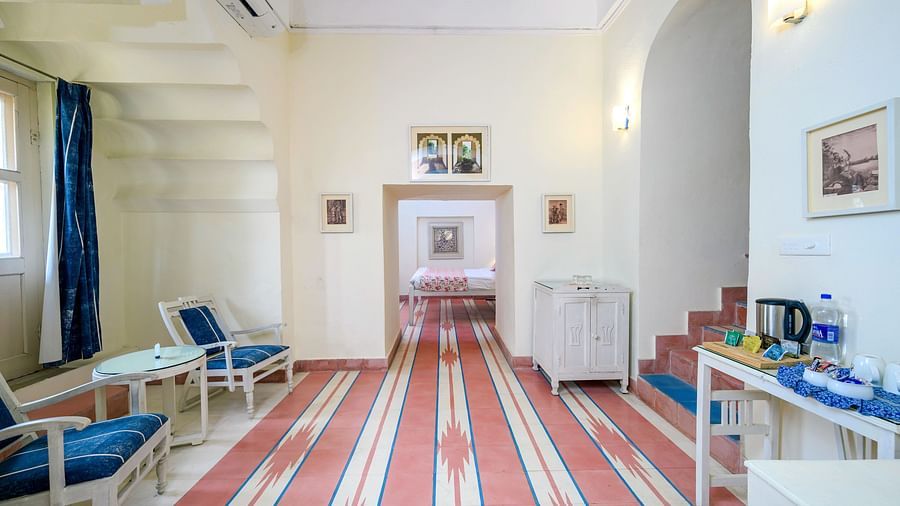 A hotel room with a seating area with chairs and a table, a refreshment station with a kettle and water bottles, and a view into another room with a bed at Tijara Fort-Palace - 19th Century, Alwar.