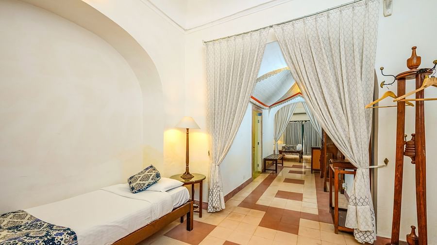 Room with a single bed, bedside lamp, clothing rack, and draped white curtains leading to another area with a bed at Tijara Fort-Palace, Alwar.
