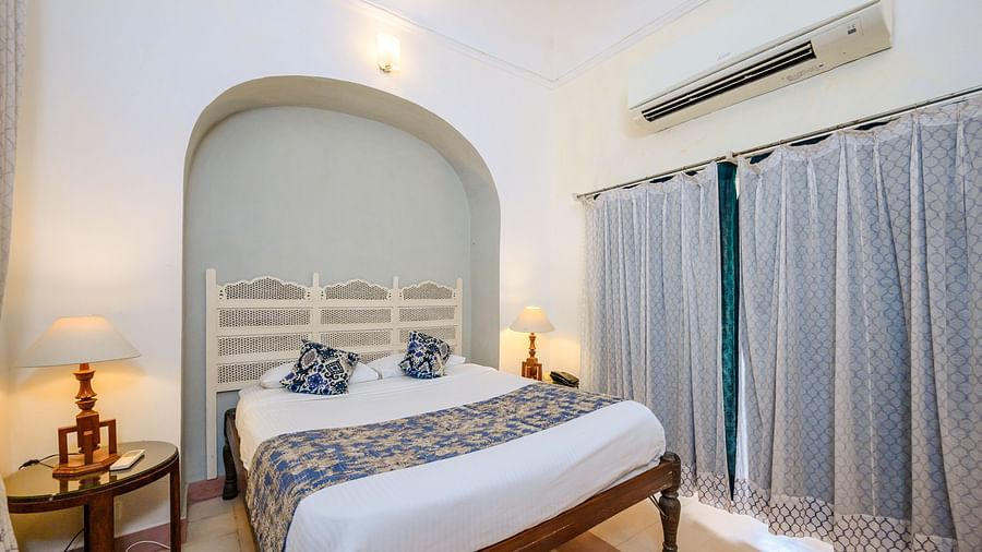 Bedroom with a bed, decorative headboard, bedside tables with lamps, curtains, and air conditioning at Tijara Fort, Alwar.