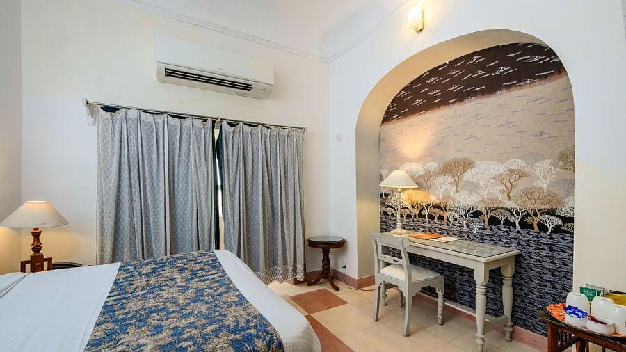 Bedroom with a bed featuring a blue and gold patterned cover, bedside lamps, a desk in an arched alcove, curtains, and AC at Tijara Fort-Palace, Alwar.