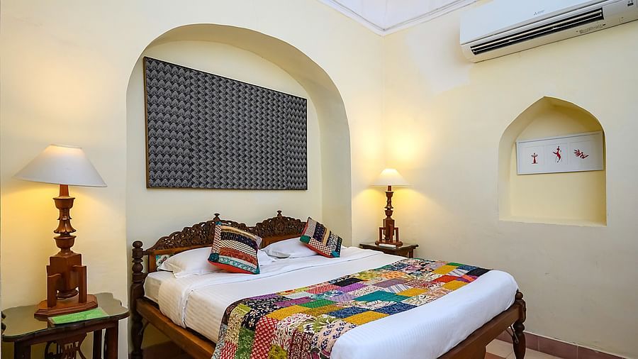 Room with bed, patterned runner and pillows, furniture including tables and lamps, arched alcoves, artwork, and wall-mounted air conditioner - Tijara Fort-Palace - 19th Century, Alwar.