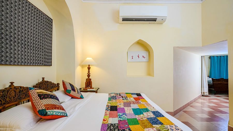 Room with bed, patterned pillows and runner, lamp, wall-mounted air conditioner, artwork, arched alcove, and doorway leading to another room with patterned floor - Tijara Fort-Palace - 19th Century, Alwar.