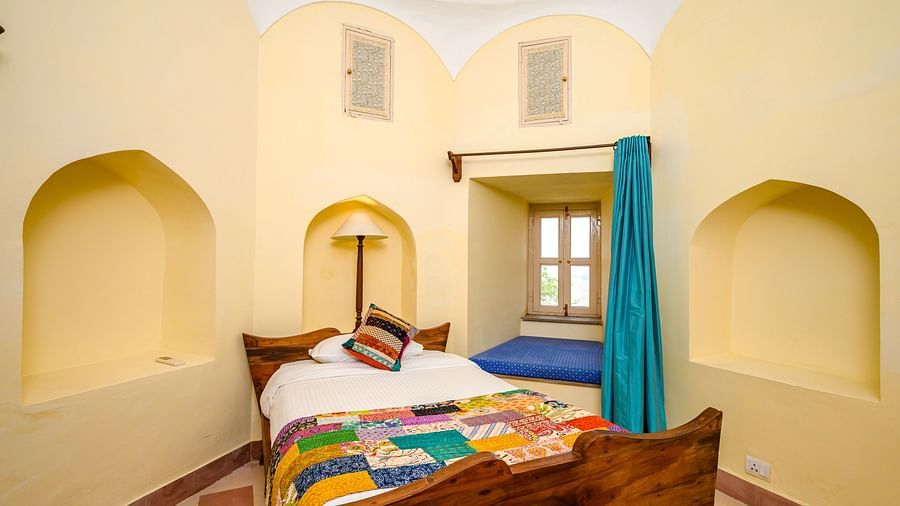 Room with bed, patterned runner and pillow, arched alcoves, window seat with cushion, lamp, and small windows - Tijara Fort-Palace - 19th Century, Alwar.