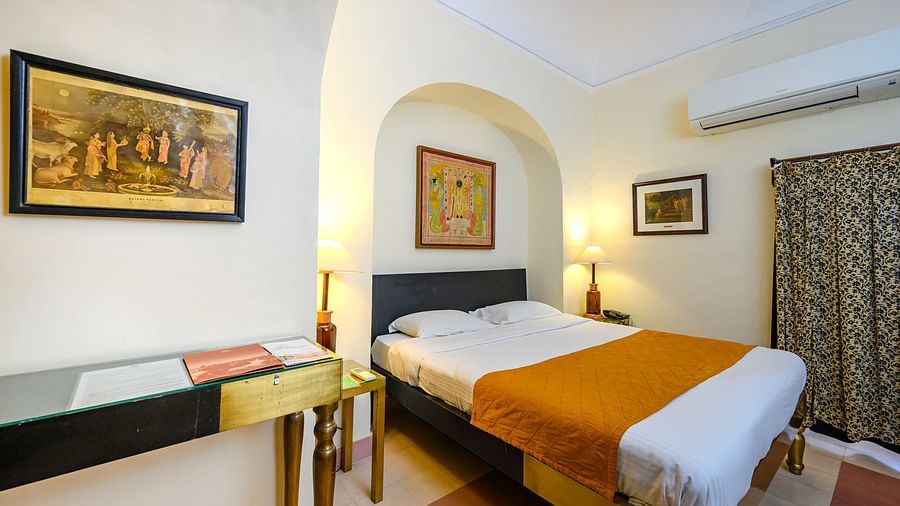 Bedroom with a bed featuring an orange runner, bedside lamps, a desk, artwork, curtains, and AC at Tijara Fort-Palace, Alwar.