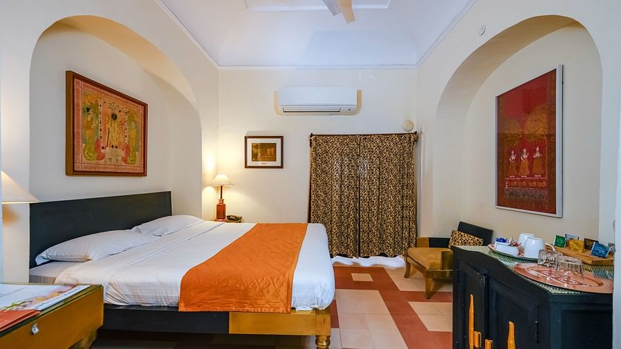 Bedroom with a bed featuring an orange runner, bedside lamps, a dark cabinet, a small seating area, artwork, curtains, and AC at Tijara Fort-Palace, Alwar.