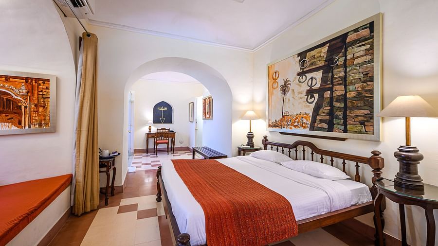 Bedroom with a bed, bedside tables with lamps, artwork, a window seat, and a view through an archway to a desk at Tijara Fort-Palace, Alwar.