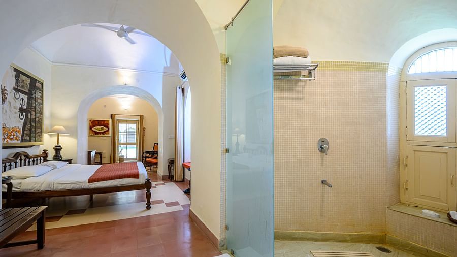 Hotel room with a bed, bedside lamps, artwork, arched doorways, and a view into a bathroom with a shower and window at Tijara Fort-Palace, Alwar.