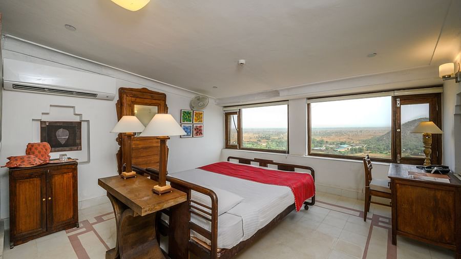 Room with bed, wooden frame, red runner, patterned floor, wooden furniture including a desk and cabinet, lamps, and windows with an outdoor view - Tijara Fort-Palace - 19th Century, Alwar.