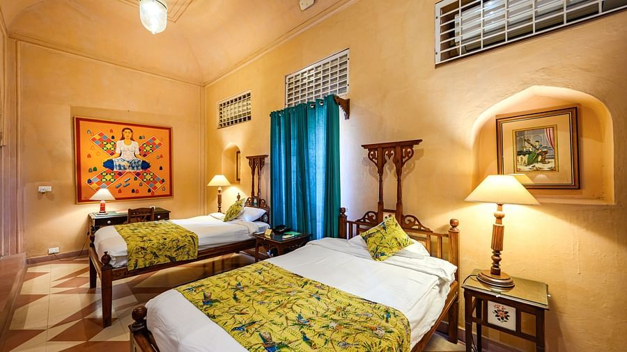A room with two beds, a table with a lamp, artwork on the wall, and a window with a grill at Tijara Fort-Palace - 19th Century, Alwar.