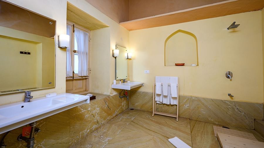 A bathroom with two sinks, two mirrors, a window, a towel rack, and a recessed arch in the wall at Tijara Fort-Palace - 19th Century, Alwar.