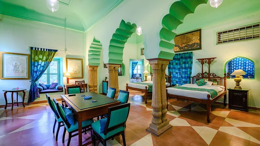 A room with a bed, a table with chairs, an arched entryway, and a window seat in Tijara Fort-Palace - 19th Century, Alwar.