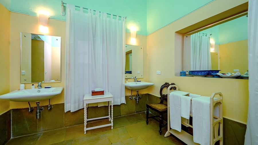 A bathroom with two sinks, mirrors, a shower curtain, a table, and a towel rack with towels in Tijara Fort-Palace - 19th Century, Alwar.