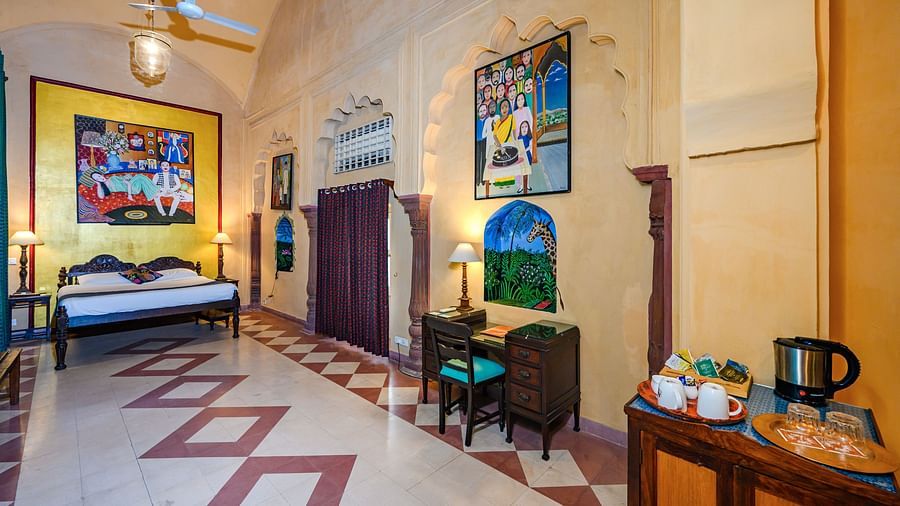 A bedroom with a bed, two side tables, two chairs, and a cabinet with a tea set in Tijara Fort-Palace - 19th Century, Alwar.