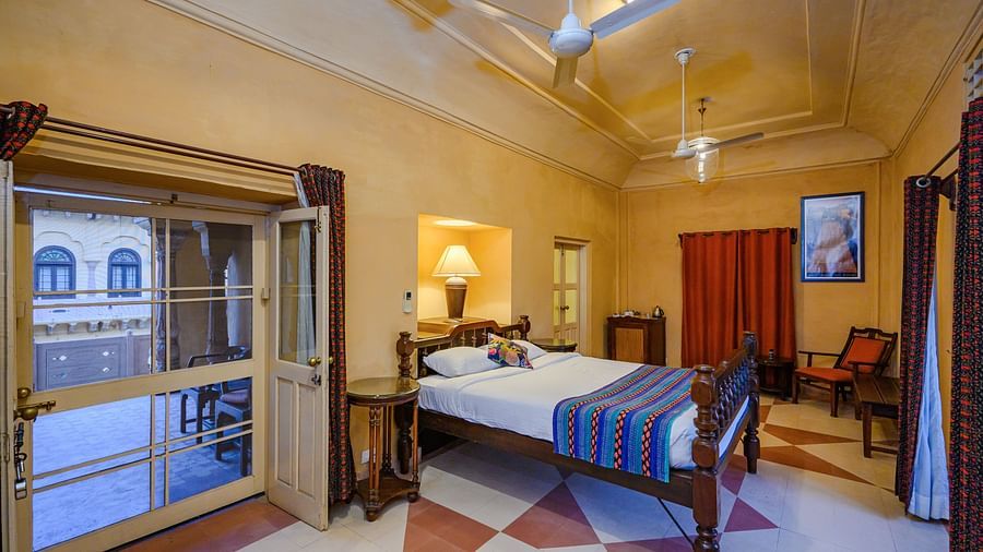 A bedroom with a bed, two side tables, two chairs, and a door opening to the outside in Tijara Fort-Palace - 19th Century, Alwar.