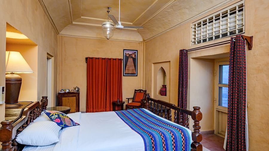 A bedroom with a bed, two chairs, a lamp, and two curtains in Tijara Fort-Palace - 19th Century, Alwar.