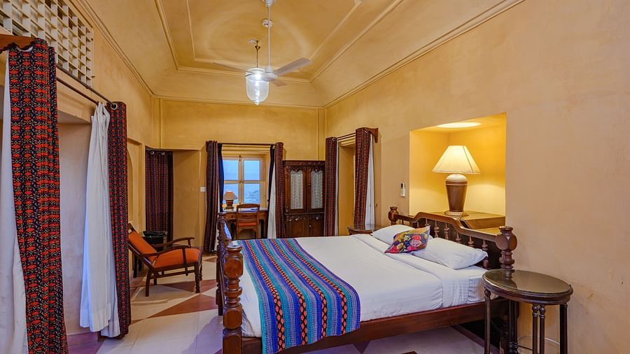 A bedroom with a bed, two side tables, a chair, and a door opening to the outside in Tijara Fort-Palace - 19th Century, Alwar.