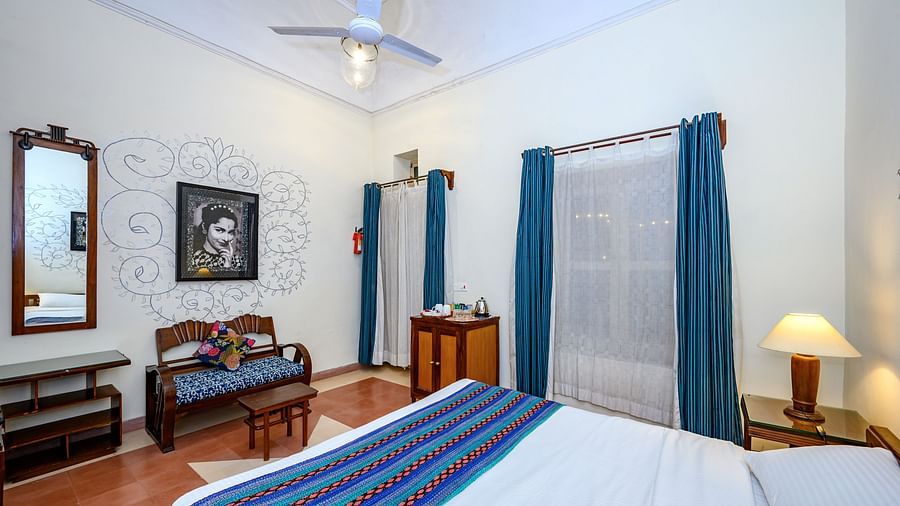 Bedroom with a bed featuring a blue and red striped runner, bedside tables with lamps, a seating area with a bench and a small table, a mirror, artwork, blue curtains, and a ceiling fan at Tijara Fort-Palace, Alwar.