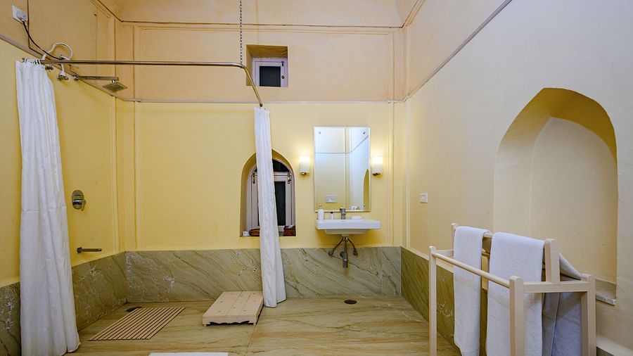 A bathroom with a sink, mirror, shower curtain, and a towel rack with towels in Tijara Fort-Palace - 19th Century, Alwar.