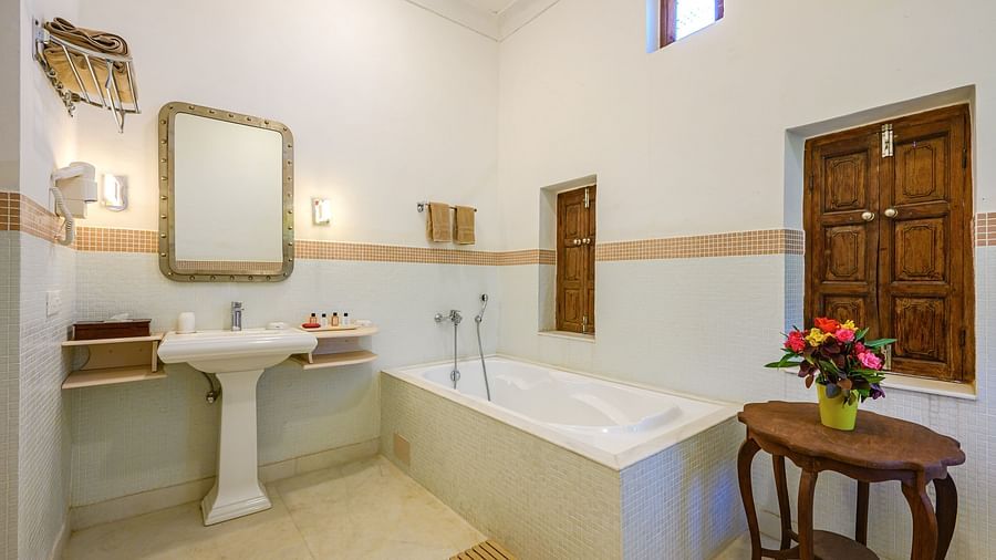 Bathroom with bathtub, sink, mirror, windows with wooden shutters, towel rack, small table with flowers, and tiled floor - Tijara Fort-Palace - 19th Century, Alwar.