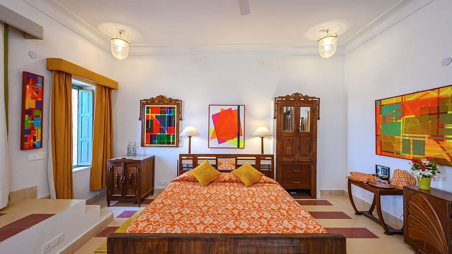 Room with bed, patterned bedding, furniture including tables, cabinets, and a wardrobe, artwork, patterned rugs, curtains, decorative ceiling lights, and patterned floor tiles - Tijara Fort-Palace - 19th Century, Alwar.