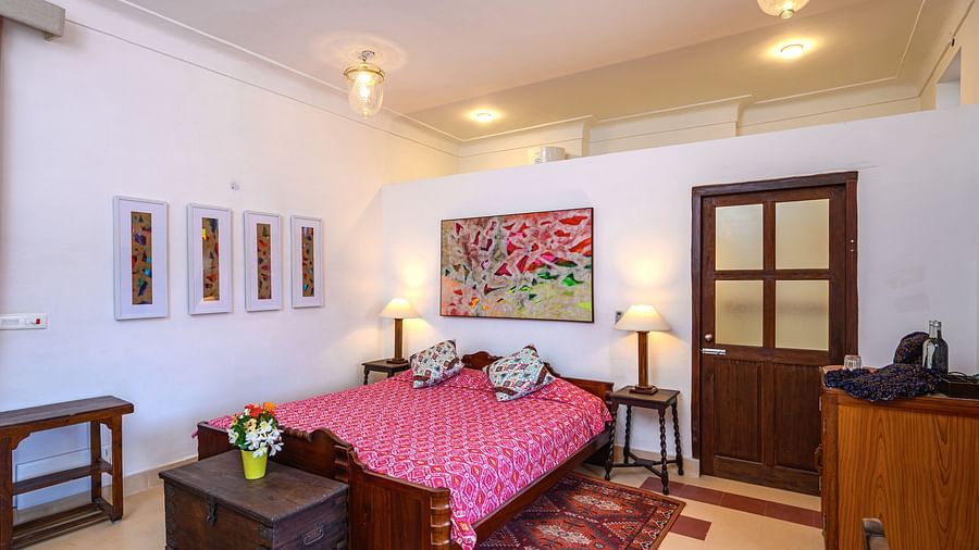 Room with bed, patterned bedding, furniture including tables, trunk, and cabinet, artwork, patterned rug, decorative ceiling lights, and patterned floor tiles - Tijara Fort-Palace - 19th Century, Alwar.